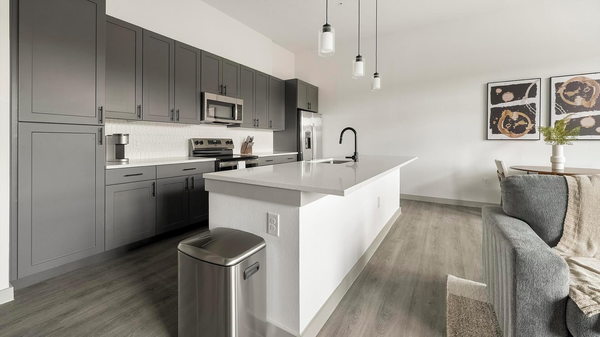 Une cuisine moderne avec des meubles gris, un îlot central blanc, des appareils en acier inoxydable et des luminaires suspendus.