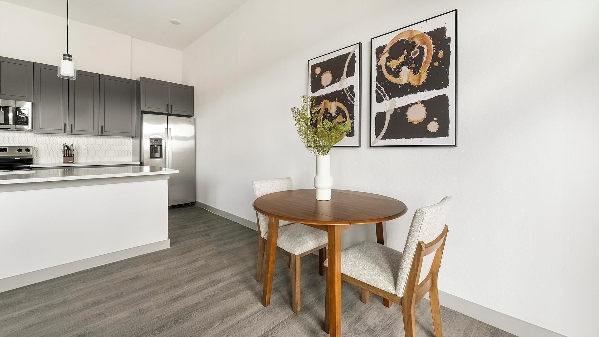 A modern kitchen with grey cabinets, a stainless steel refrigerator, and a white countertop. There is a dining area with a wooden table and two chairs, and a vase with green foliage on the table.