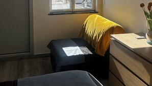 Een slaapkamer met een raam, een bed, een stoel, een dressoir en een vaas met bloemen.