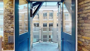 A narrow, enclosed elevator shaft with metal mesh doors, leading to a building with multiple windows and a brick facade. - Stylish Penthouse with City Views and Aircon - Pass the Keys (London)