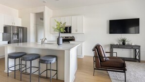A modern kitchen with white cabinets, a large island with bar stools, a microwave, and a TV mounted on the wall. - Landing | Modern 2BD, Pool (Casas Adobes)