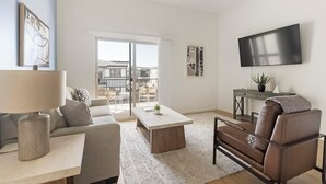 Een moderne woonkamer met een bank, salontafel en een leren fauteuil. Een televisie is aan de muur bevestigd en er is een balkon met uitzicht.
