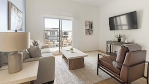 A modern living room with a sofa, coffee table, and leather armchair. A TV is mounted on the wall, and there's a balcony with a view. - Landing | Stunning 2BD, Pool (Casas Adobes)