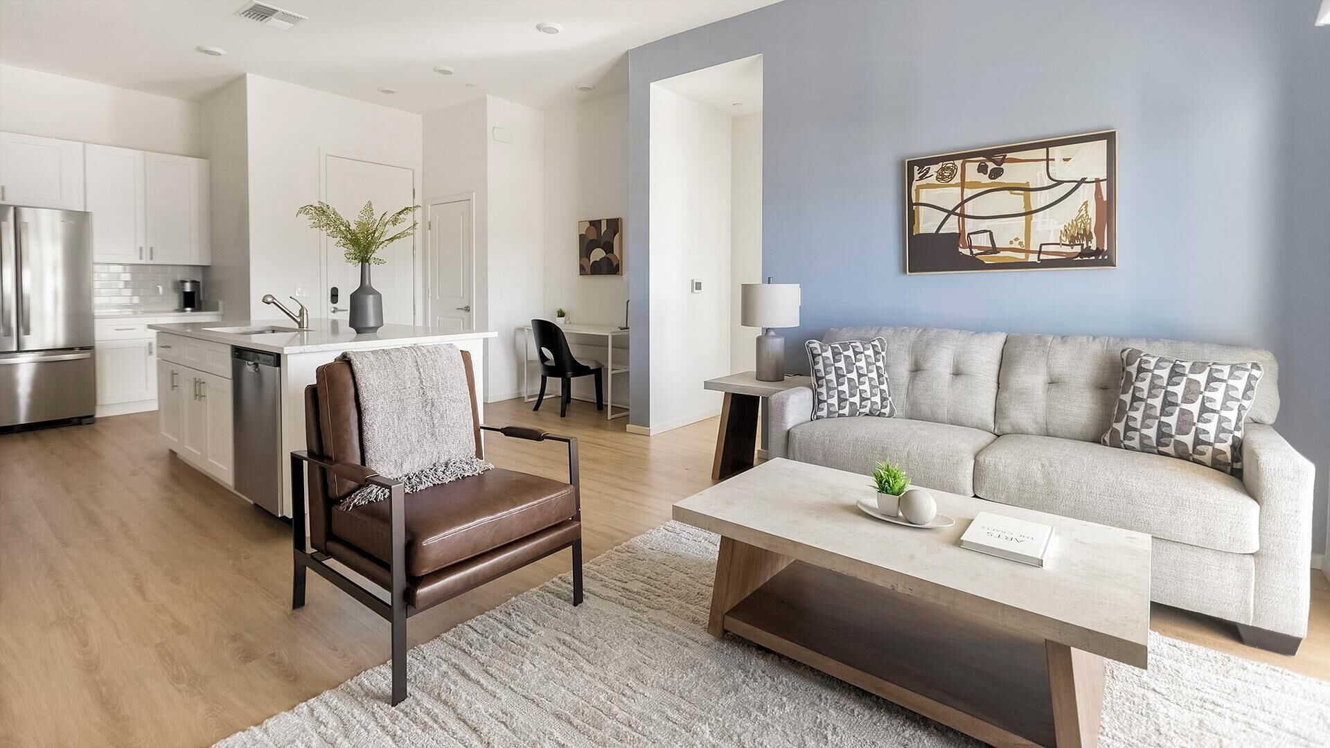 Ein modernes Wohnzimmer mit einer Couch, einem Couchtisch und einem Sessel. Im Hintergrund ist eine Küchenzeile mit Schränken und einem Kühlschrank zu sehen.