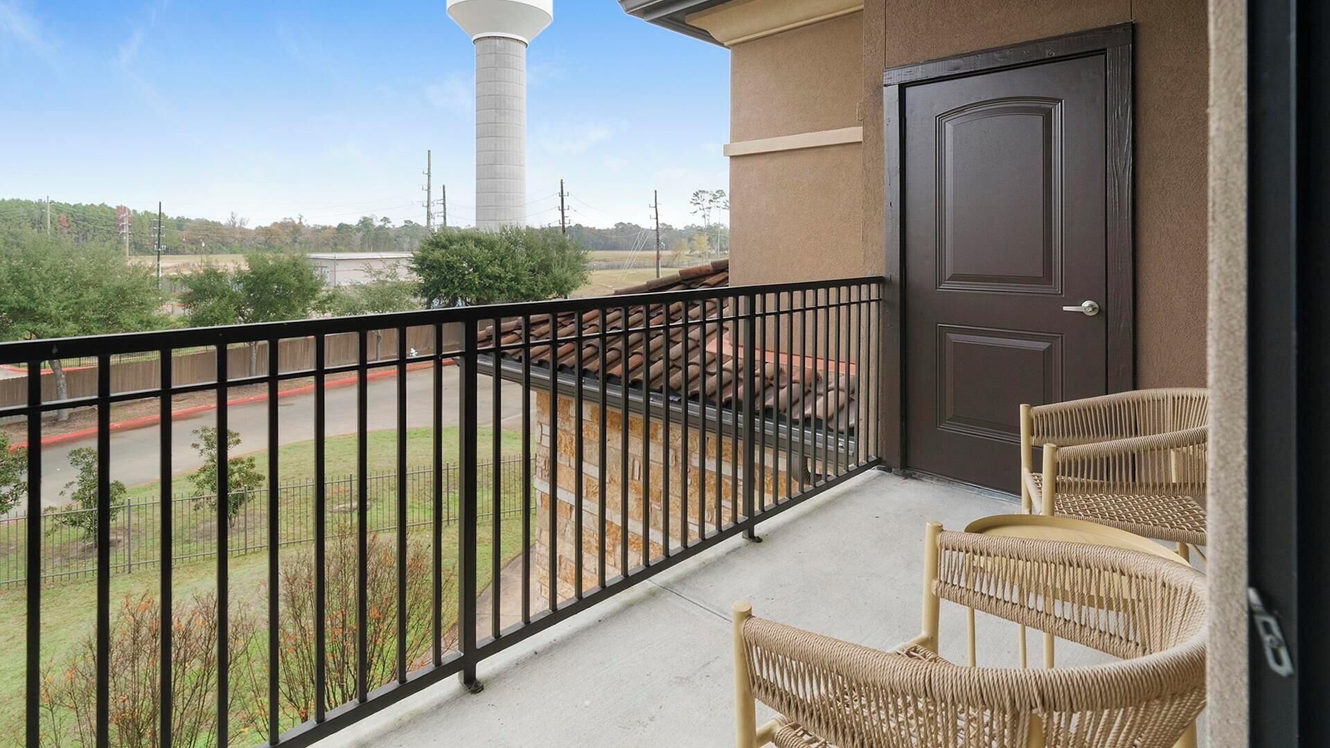 Un balcone con sedie in vimini, una porta e vista su un parco e una torre d'acqua.