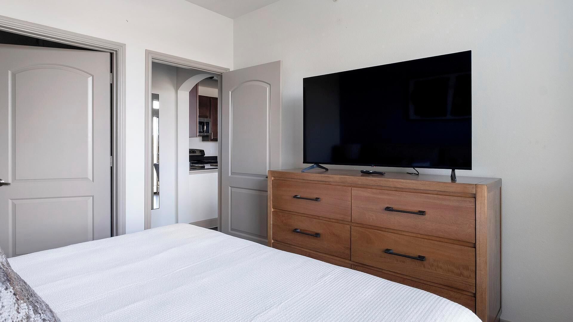 A bedroom with a bed, a wooden dresser, a television on the wall, and a door leading to a bathroom.