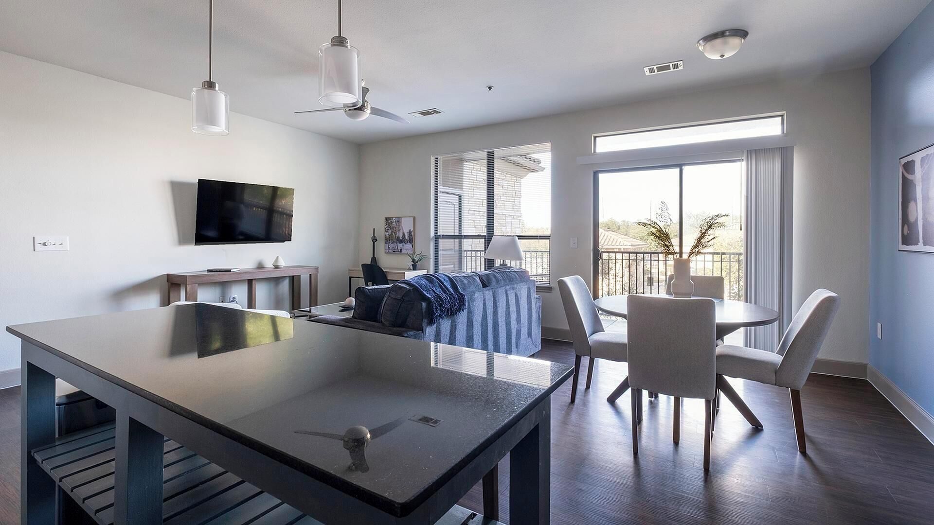 A modern living room with a large flat-screen TV, a dining area with a round table and chairs, and a balcony with a view.