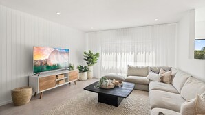 Ein modernes Wohnzimmer mit Flachbildfernseher, einer Couch, einem Couchtisch und einem großen Fenster mit transparenten Vorhängen.
