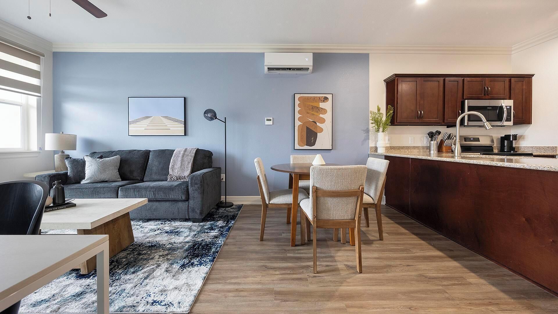 Un salon moderne avec un canapé gris, une table à manger avec des chaises, un coin cuisine doté de meubles en bois et d’un four à micro-ondes, et un mur bleu orné d’œuvres d’art.