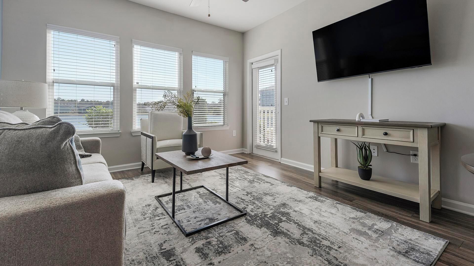 Un salon avec un canapé gris, une petite table sur laquelle est posé un vase, un téléviseur fixé au mur et un bureau en bois doté de tiroirs.