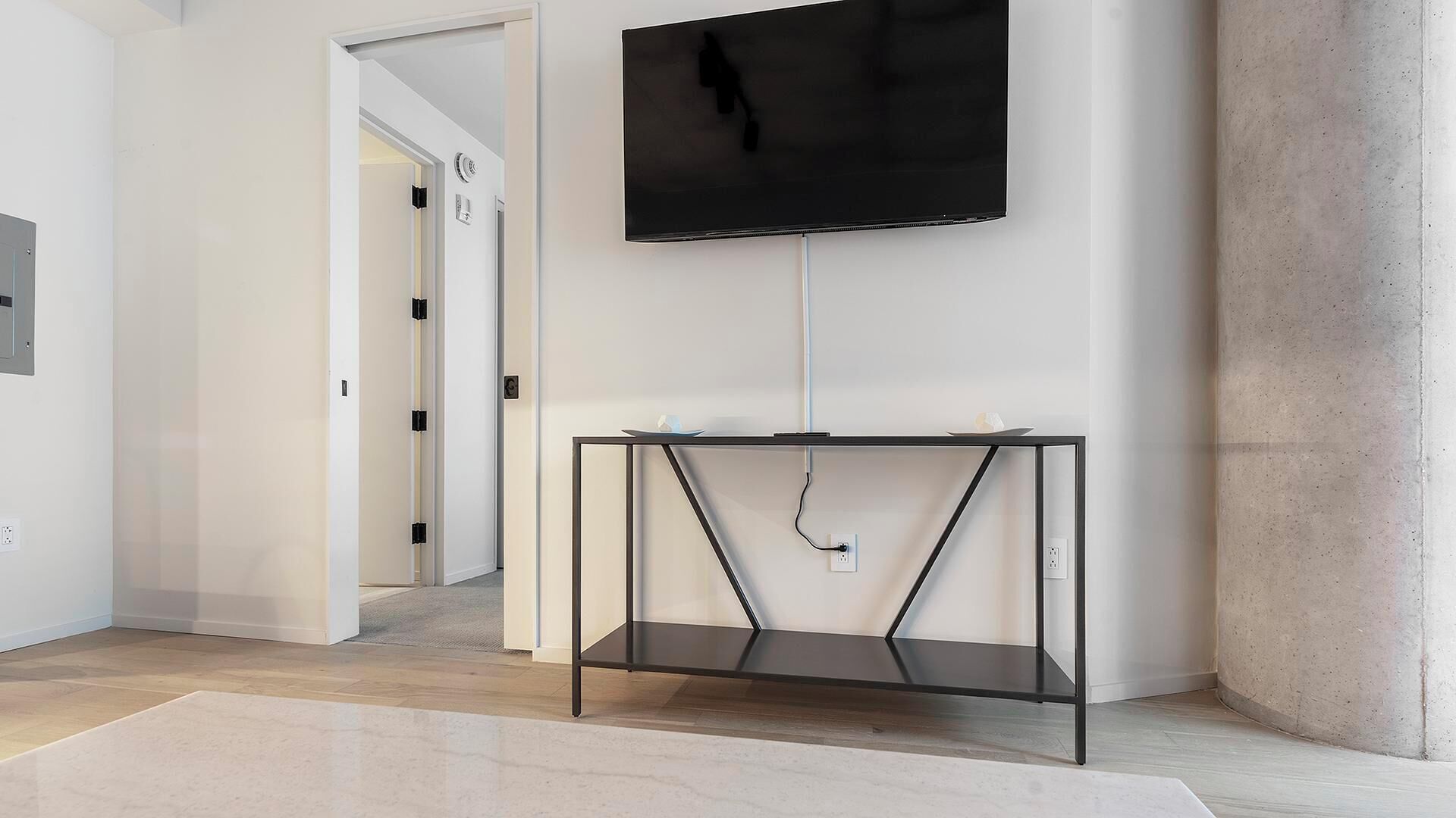 A modern, minimalist room with a flat-screen TV mounted on the wall, a black metal console table, and a light-colored carpet.