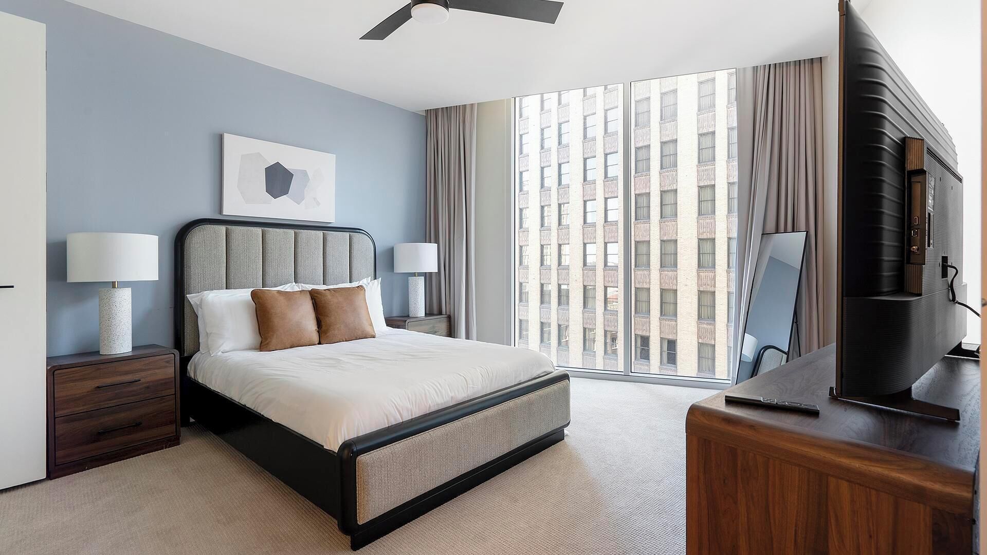 A modern hotel room with a large bed, two bedside tables with lamps, a flat-screen TV, and a view of a building through large windows.