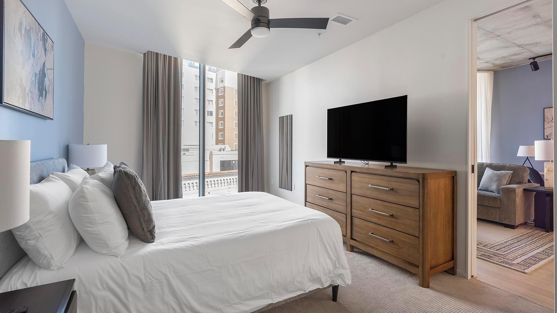 A hotel room with a bed, a TV, a wooden dresser, and a view of buildings through the window.