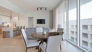 Moderni hotellihuone, jossa on ruokailutila, keittiö ja suuri ikkuna, josta avautuu näkymä kaupunkiin.