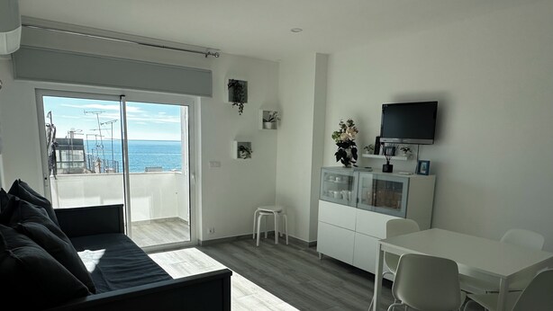 Uma sala de estar moderna com sofá, uma mesa de jantar com cadeiras, área de cozinha com televisão e uma porta de vidro deslizante que dá para uma varanda com vista para o mar.