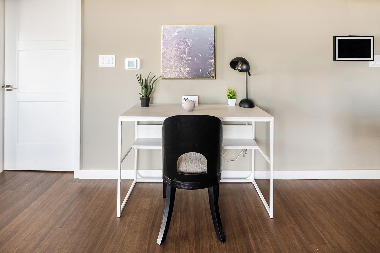 A room with a white desk, a black chair, a framed picture, a floor lamp, and a small potted plant.