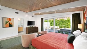 A hotel room with a wooden ceiling, a large bed with a red quilt, a television on a wooden dresser, a small round table, and a view of a garden through a sliding glass door. - Central Palm Springs | Pool | Hot Tub (Palm Springs)
