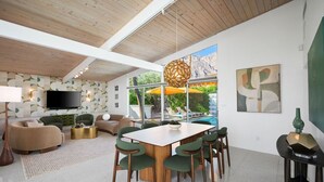 A modern living room with a wooden ceiling, a TV mounted on the wall, a pool visible through large windows, and a dining area with green chairs. - Central Palm Springs | Pool | Hot Tub (Palm Springs)