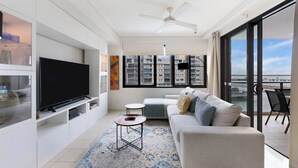 A modern living room with a flat-screen TV, a sofa, a coffee table, and a large window offering a city view. - Lagoon Panorama Relaxed Waterfront Escape near CBD (Darwin)