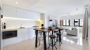 A modern kitchen with white cabinetry, a central island with stools, and a dining area with a sofa and large windows. - Lagoon Panorama Relaxed Waterfront Escape near CBD (Darwin)