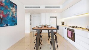 A modern kitchen with white cabinets, a dining area with a wooden table and chairs, and a large abstract painting on the wall. - Lagoon Panorama Relaxed Waterfront Escape near CBD (Darwin)