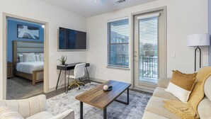 Un salon moderne comprenant un lit, un bureau et un canapé. On y trouve une grande fenêtre avec des stores, un téléviseur à écran plat fixé au mur et une lampe sur un pied.