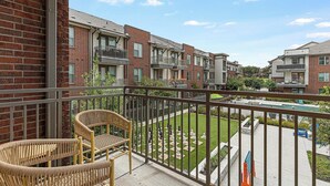 Un balcon avec des fauteuils en osier, une vue sur un quartier résidentiel avec des immeubles en briques et un petit jardin avec des pièces d’échecs.