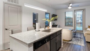 Une cuisine moderne dotée de plans de travail en granit, d’un lave-vaisselle et d’un four encastré. On y trouve un coin repas avec une table et des chaises, ainsi qu’un salon avec un canapé et un tapis.