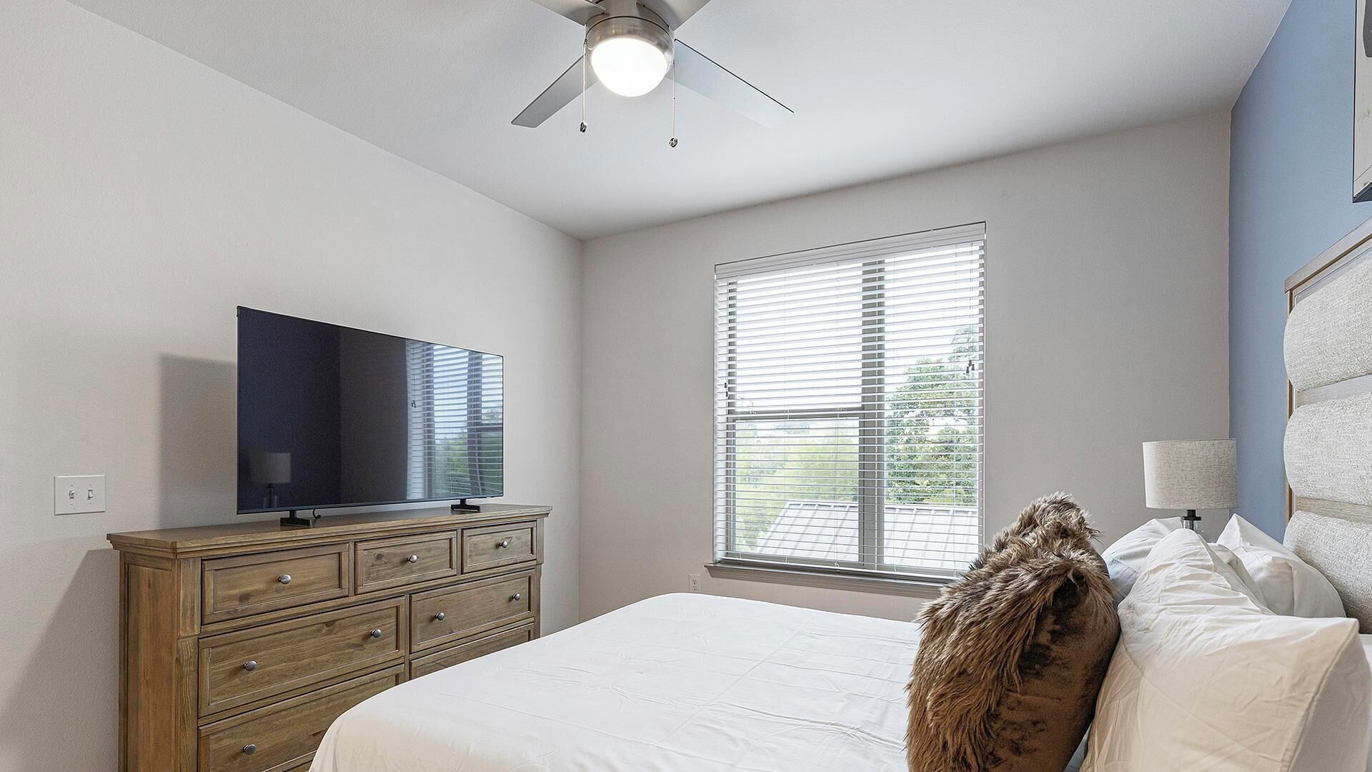 A bedroom with a bed, a dresser, a television, a ceiling fan, and a window with blinds.