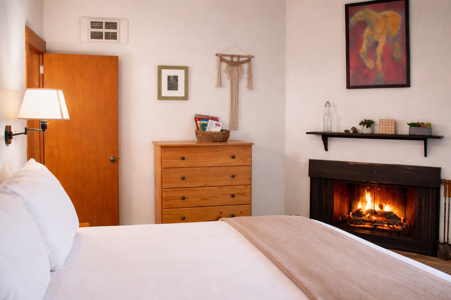 A bedroom with a bed, a wooden dresser, a wall-mounted shelf with decorative items, a framed picture, and a fireplace with a fire.
