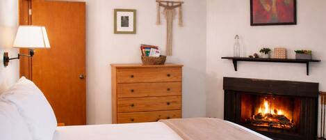 A bedroom with a bed, a wooden dresser, a wall-mounted shelf with decorative items, a framed picture, and a fireplace with a fire.