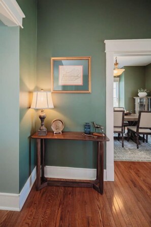 Una habitación con una consola de madera, una lámpara, un mapa enmarcado y un área de comedor al fondo.