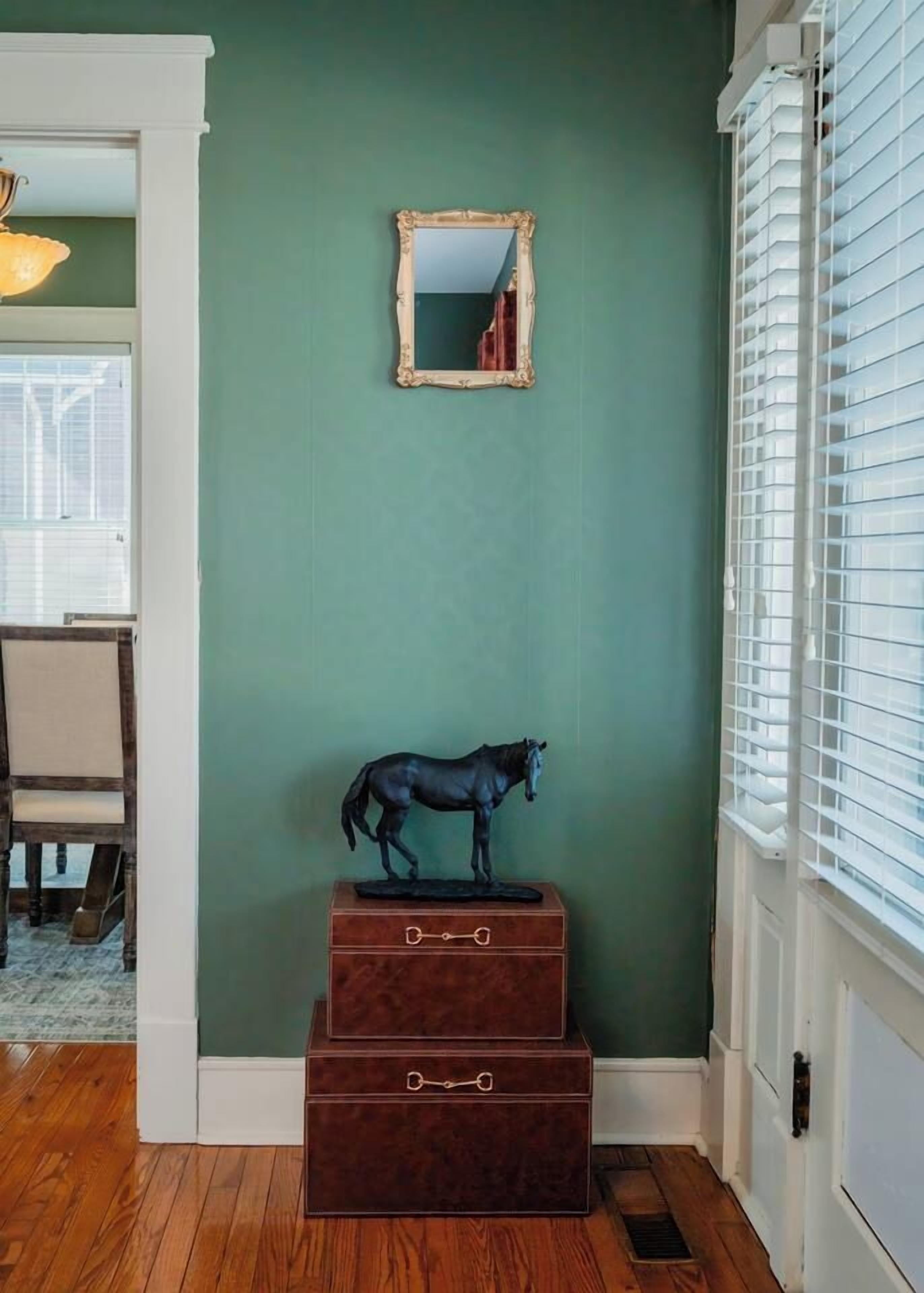 A room with a green wall, a mirror, a wooden chest of drawers, and a horse statue.