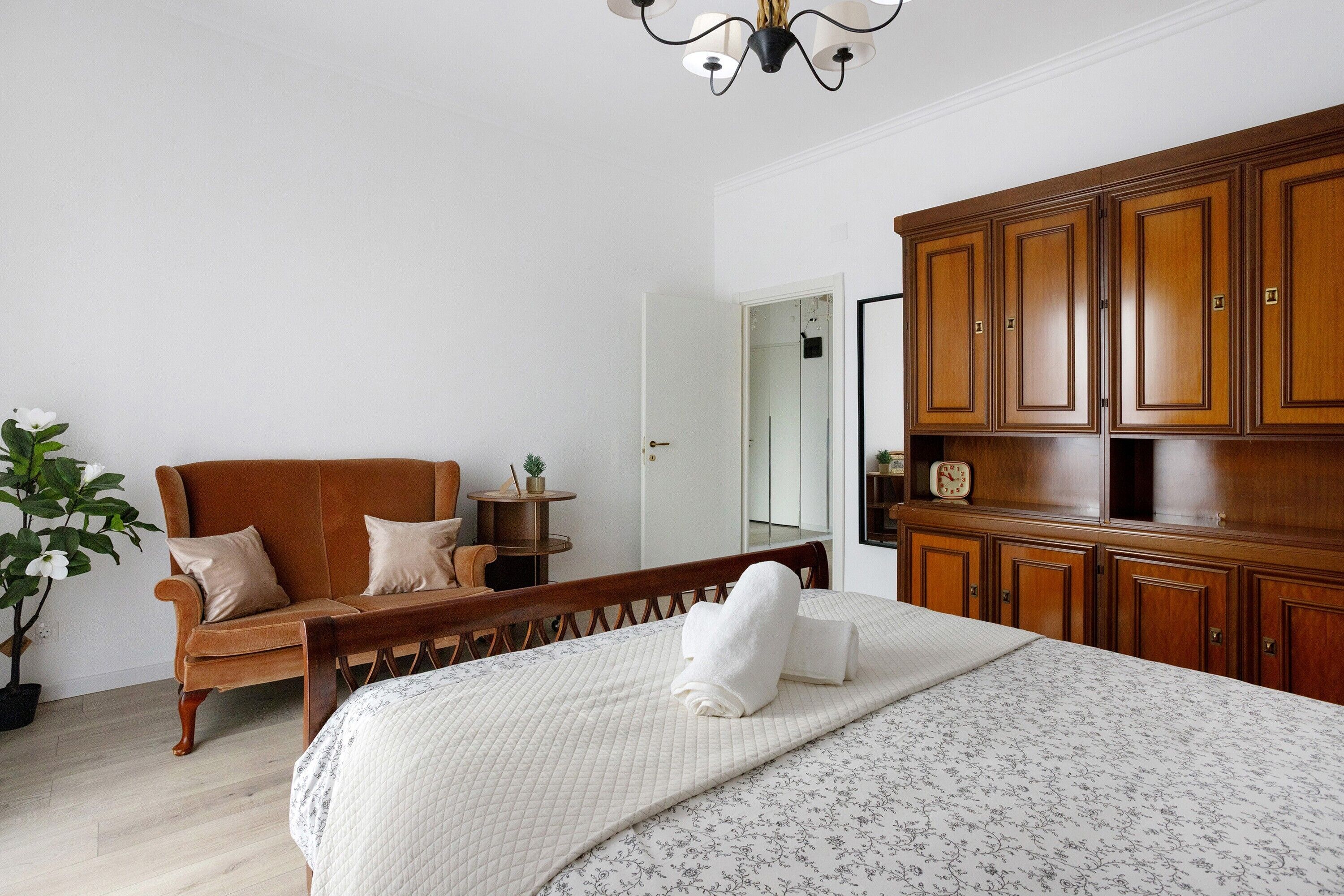 A bedroom with a bed, wooden wardrobe, a bench, a potted plant, and a mirror.