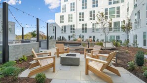 An outdoor patio with wooden furniture, a fire pit, and a view of a modern building with large windows. - Landing | Stylish 1BD, Yoga Studio, Gym (Durham)