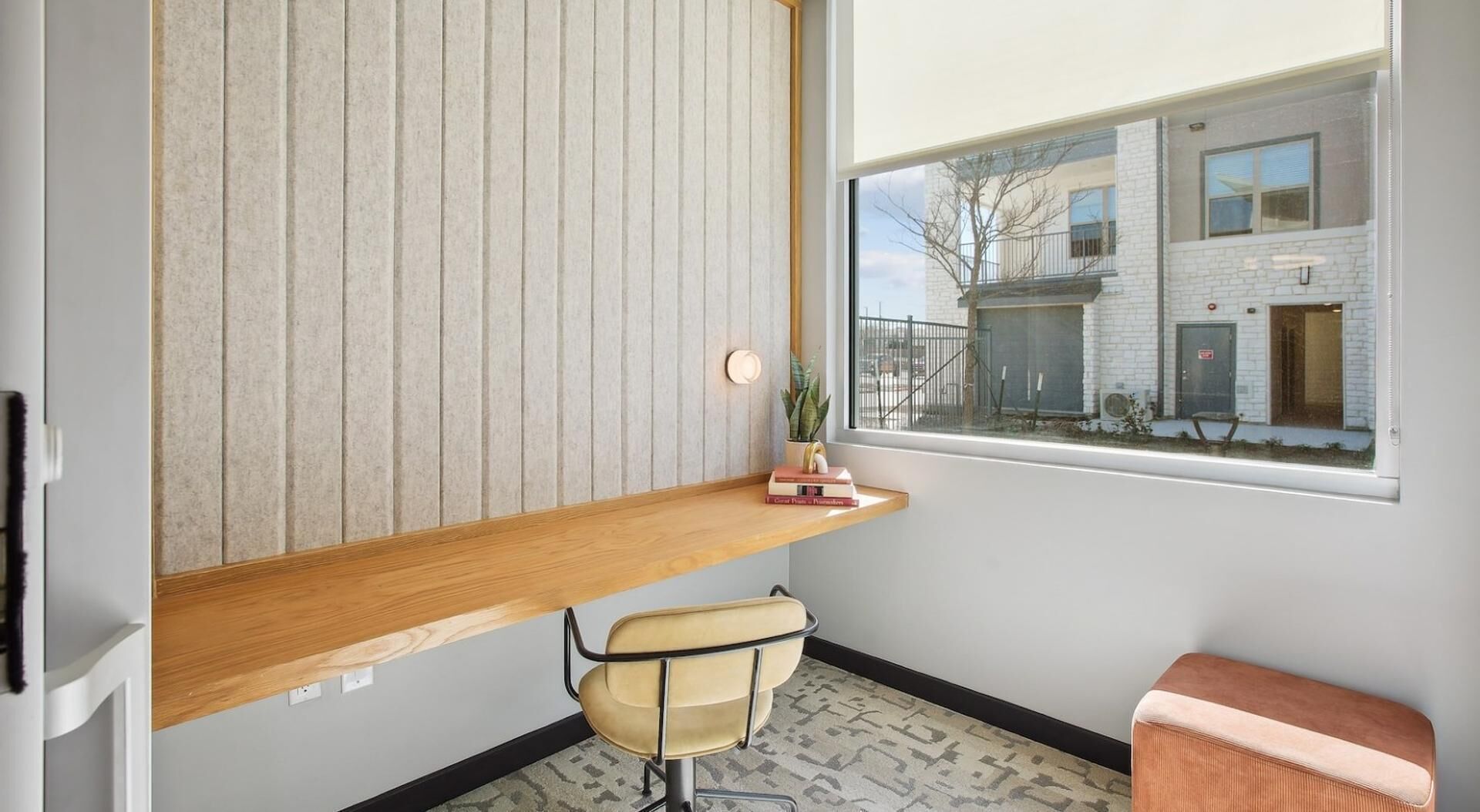 Een kleine, moderne kamer met een houten bureau, een stoel, een tafeltje met boeken en een raam met uitzicht op een gebouw.