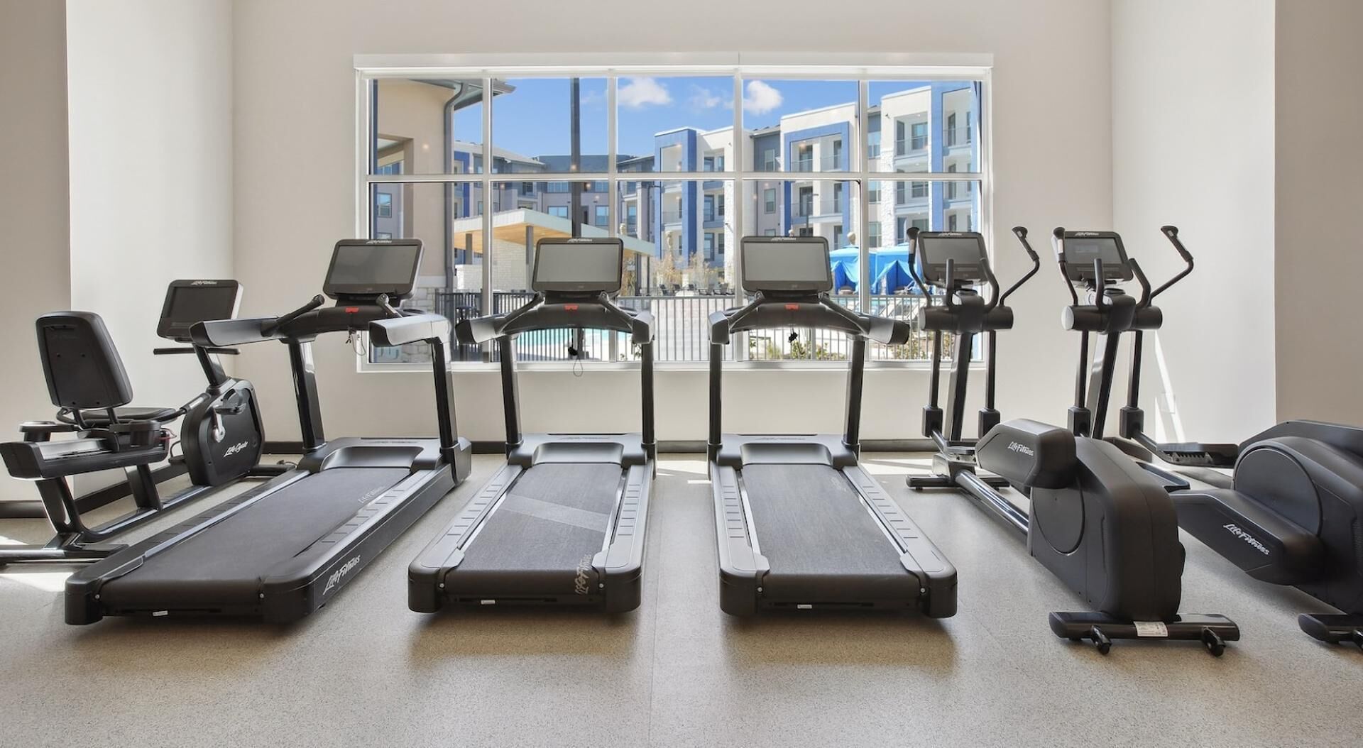A gym with treadmills, elliptical machines, and stationary bikes, with large windows offering a view of buildings outside.