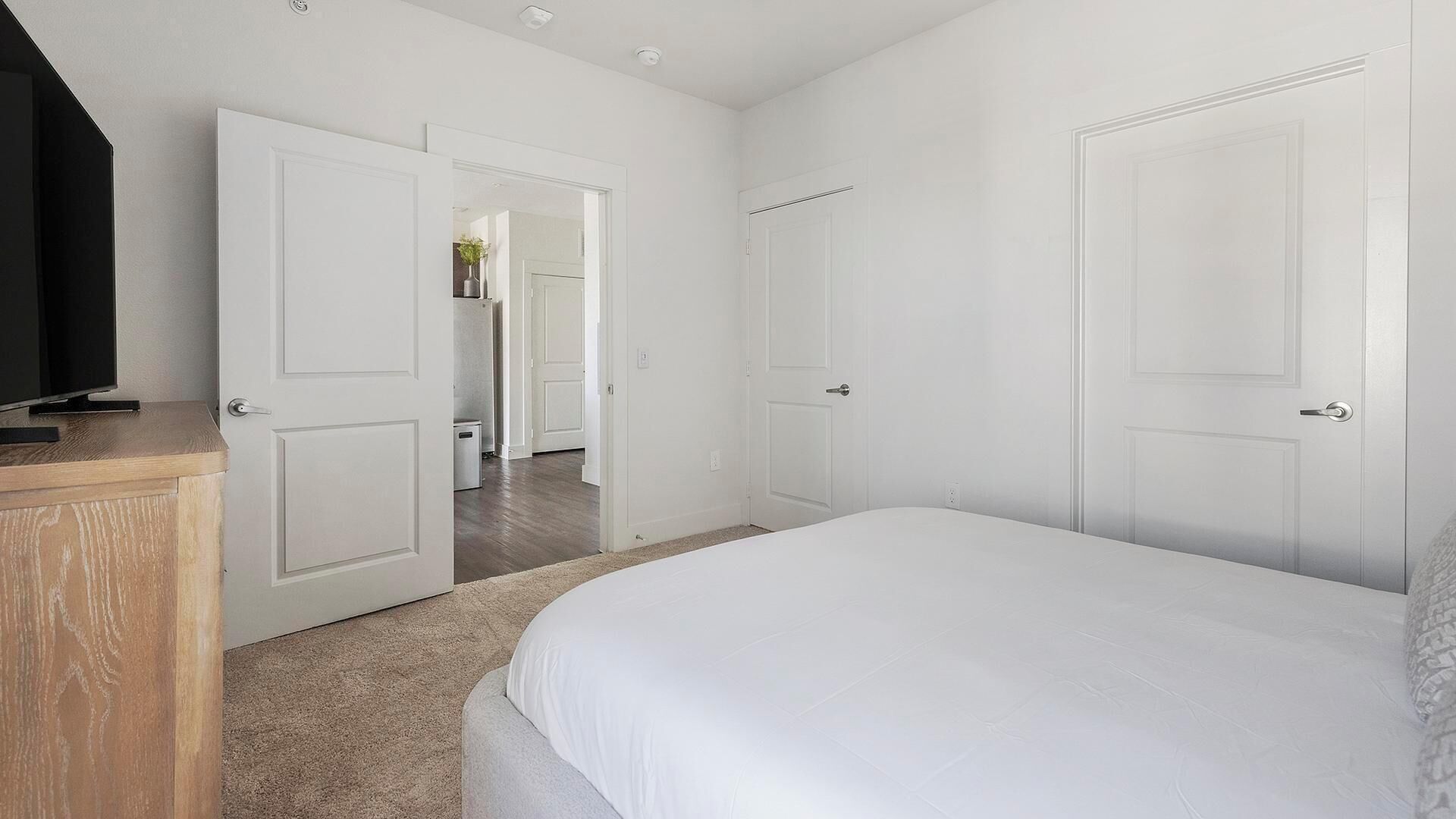 A bedroom with a bed, a TV on a wooden cabinet, and white doors leading to other rooms.