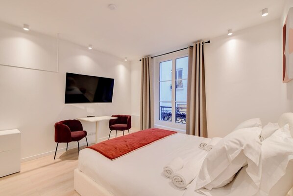 A modern hotel room with a large bed, a desk, two chairs, a TV mounted on the wall, and a window with curtains.