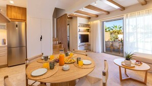 A modern dining area with a round wooden table set for breakfast, a kitchen with a built-in refrigerator, and a view of the outdoors through a sliding glass door. - Villa Kamelia Seaside Almyra by Villa Plus (Agkisaras)