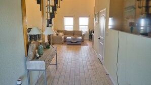 Un vaste espace de vie comprenant un canapé, une table basse et un miroir. De grandes fenêtres laissent entrer la lumière naturelle et un ventilateur de plafond est installé.