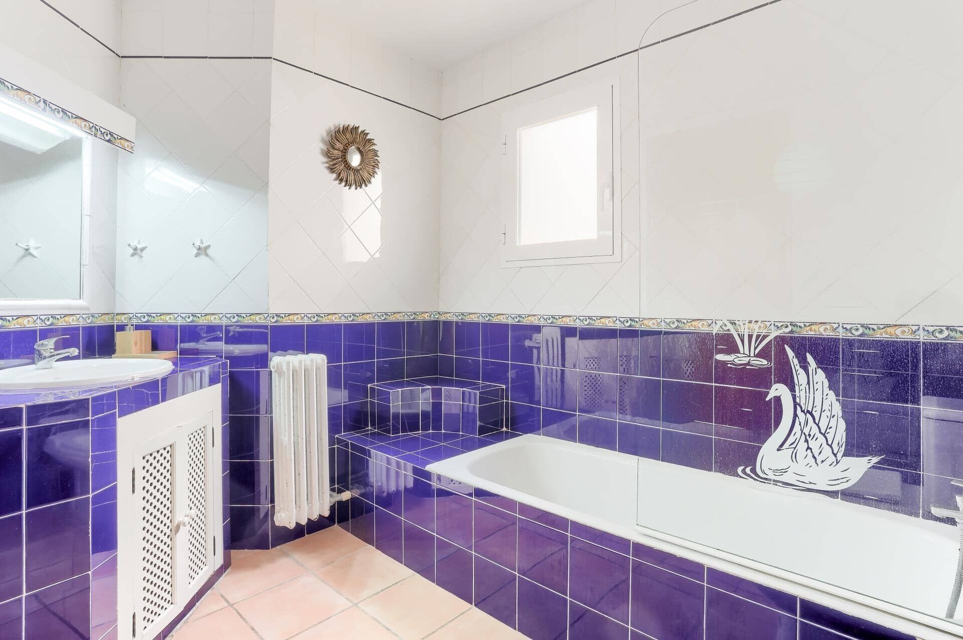 A bathroom with a white bathtub, a white sink, and a mirror. There is a decorative wreath on the wall and a window above the bathtub.