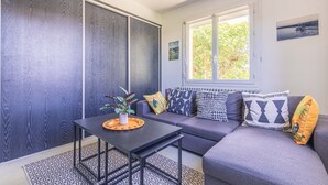 A modern living room with a sectional sofa, a small table with a plant, and a window with a view of greenery. - Apartment in Saint-Pierre-d’Oléron with Private Terrace, Garden, and Wi-Fi (Saint-Pierre-d'Oléron)