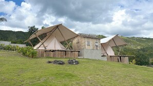 Un chalet rustique et écologique, doté de deux grandes tentes et d’une cabane en bois, niché au pied de collines verdoyantes, sous un ciel partiellement nuageux.