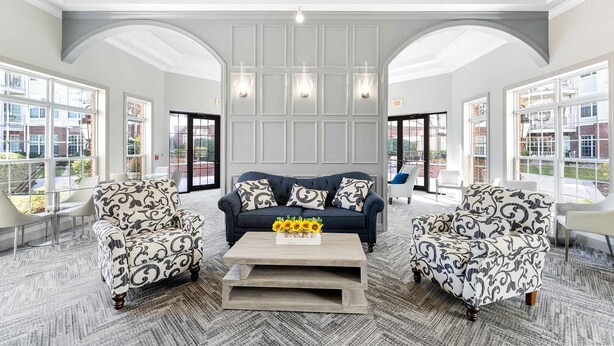 A spacious lobby with a central seating area featuring a dark blue sofa and patterned armchairs, a wooden coffee table with sunflowers, and large windows offering views of the surrounding area. - Landing | Spectacular 1BD, Pool, Courtyard (Raritan)
