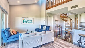 A modern living room with a blue sofa, a grey sofa, and a glass coffee table. There is a wooden staircase leading to an upper level. - The Intercoastal #209 - Intercoastal Escape (Indian Shores)
