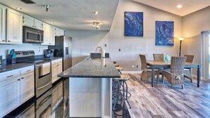 A modern kitchen with stainless steel appliances, a central island, and a dining area with wicker chairs and a wooden table. - The Intercoastal #209 - Intercoastal Escape (Indian Shores)