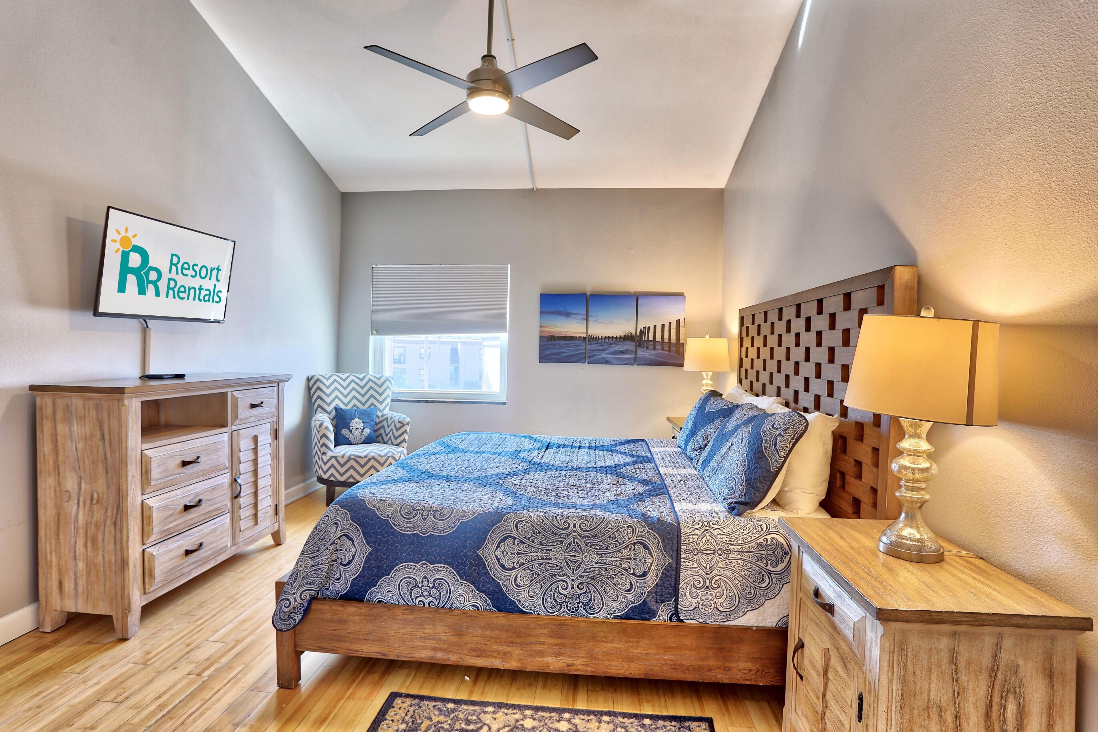 Une chambre avec un lit, une table de chevet en bois, un téléviseur affichant une page consacrée aux « locations de vacances », un ventilateur de plafond et une fenêtre avec des stores.