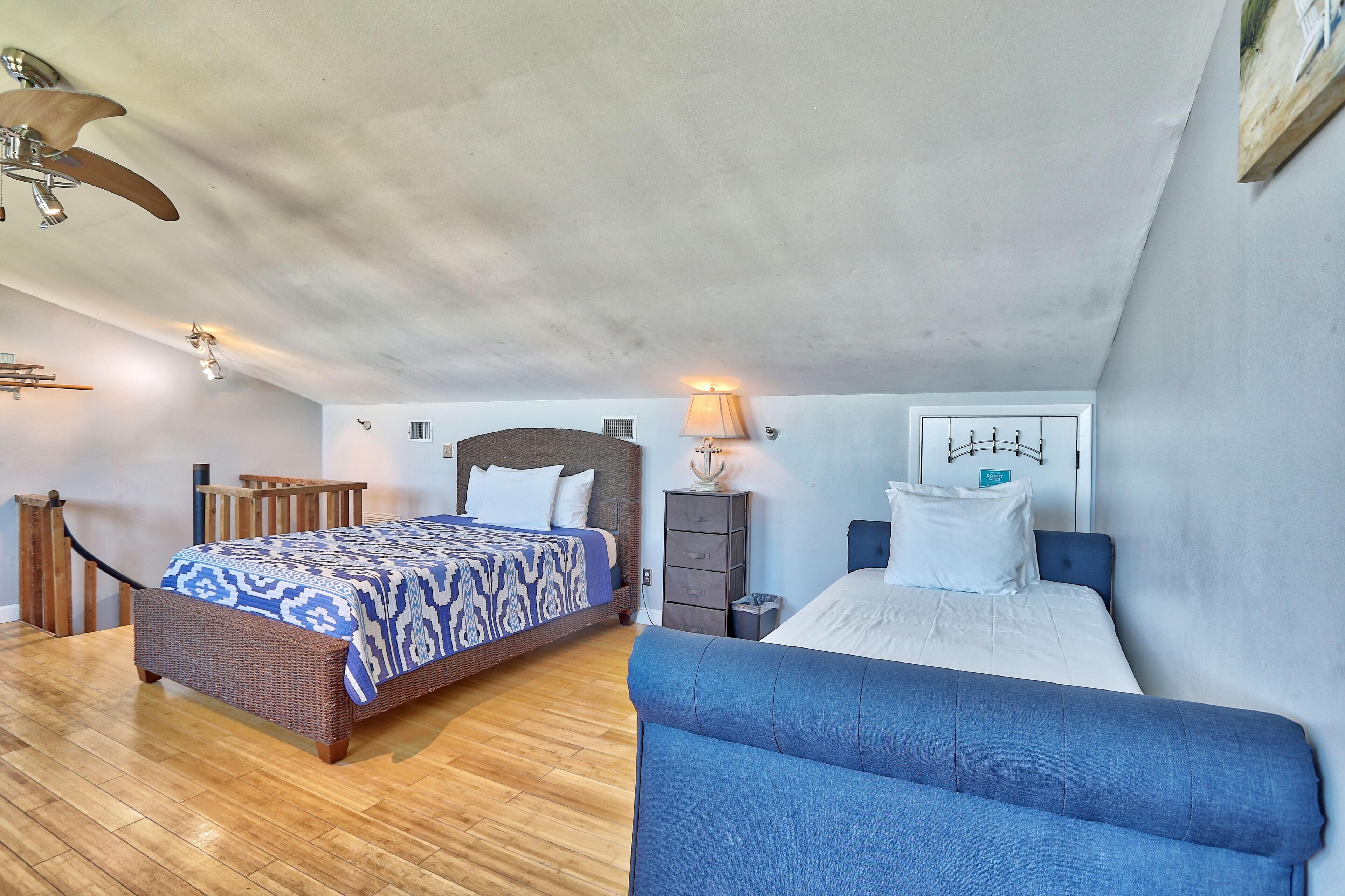 Ein Schlafzimmer mit einem großen Bett, einem kleineren Bett, einem blauen Sofa, Holzboden, Deckenventilator und einer Wandlampe.