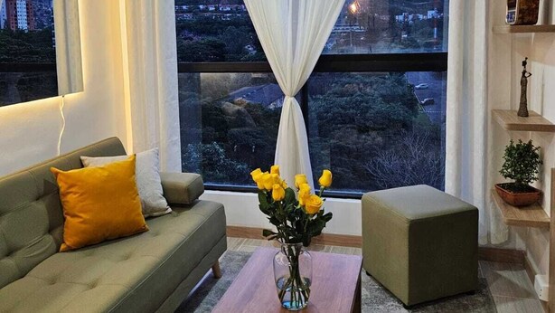 Una sala de estar con sofá, una mesa de centro y vistas a un paisaje urbano a través de amplios ventanales.